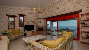Een ruime woonkamer met stenen muren, een open haard, een televisie en een balkon met uitzicht op zee.