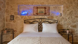 Een slaapkamer met een stenen muur, een bed met witte lakens, een lijst met een afbeelding boven het bed, en een klein tafeltje met een vaas ernaast.