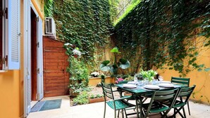 Ein Essbereich mit Tisch für vier Personen, umgeben von grünen Stühlen, in einem Garten mit Backsteinmauer und efeuüberwachsenen Wänden.