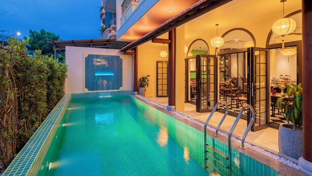 An indoor swimming pool with a tiled edge, a covered patio area with a dining table and chairs, and a view of the surrounding buildings. - BACKHOME BOUTIQUE OLD TOWN HOI AN (Da Nang)