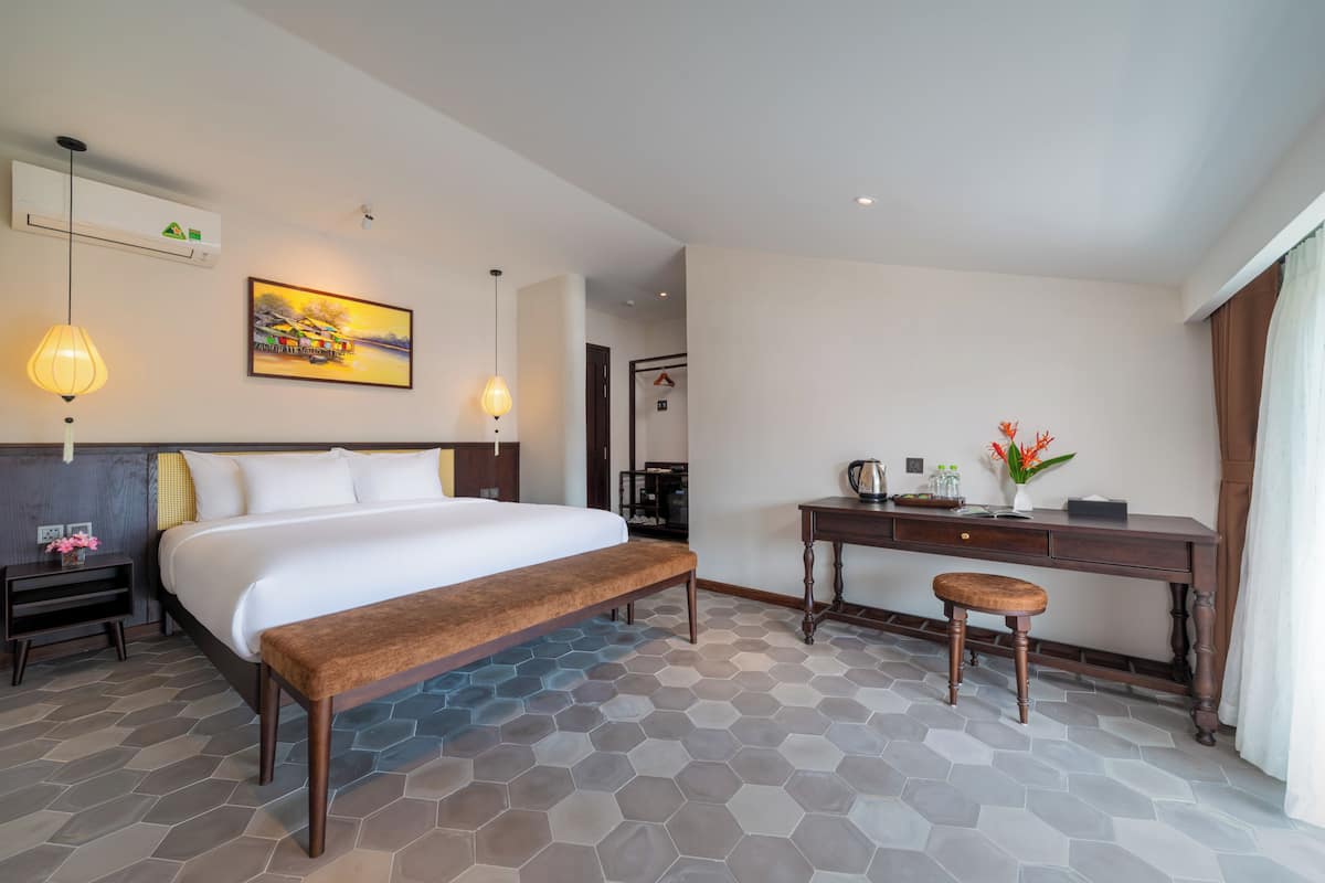 A hotel room with a bed, bench, desk, and a painting on the wall.