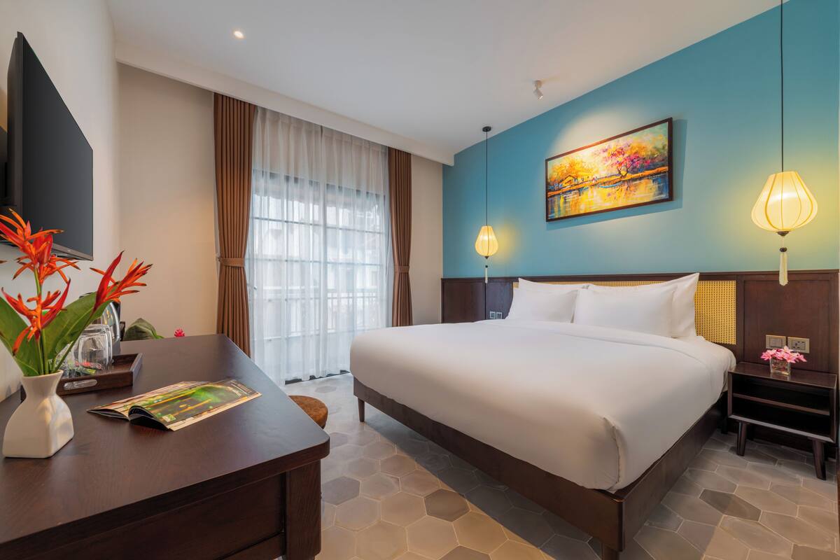 A hotel room with a large bed, a desk with a vase of flowers, a painting on the wall, and a window with curtains.