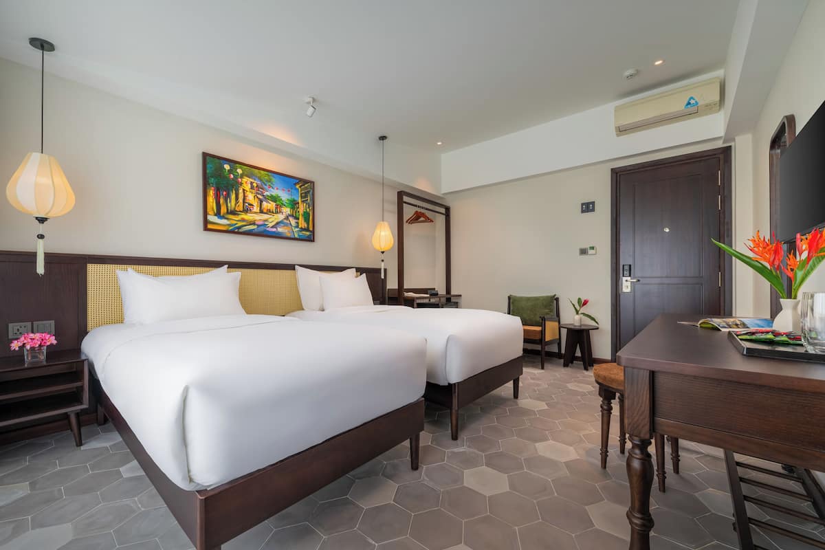 A hotel room with two beds, a desk, a chair, a painting, a mirror, and a small table with flowers.