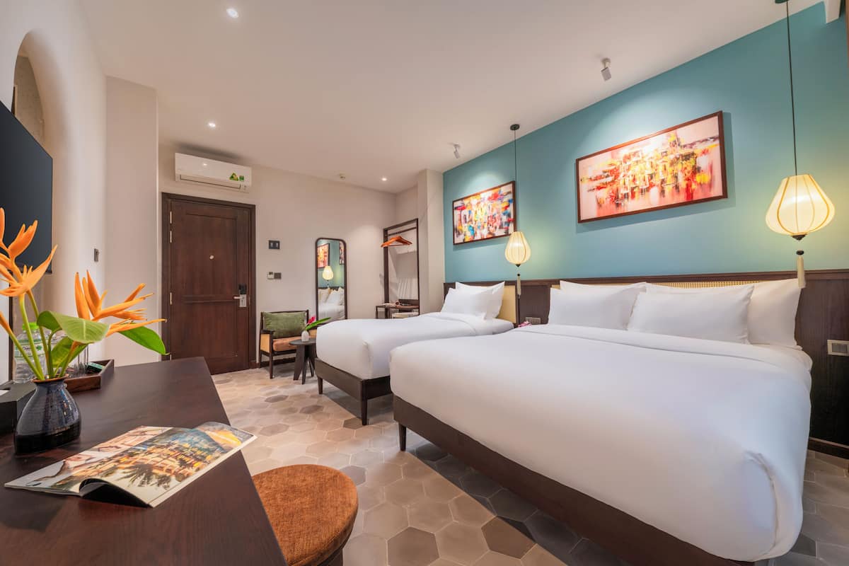 A hotel room with two beds, a desk, a chair, a vase with flowers, and two wall-mounted lamps.