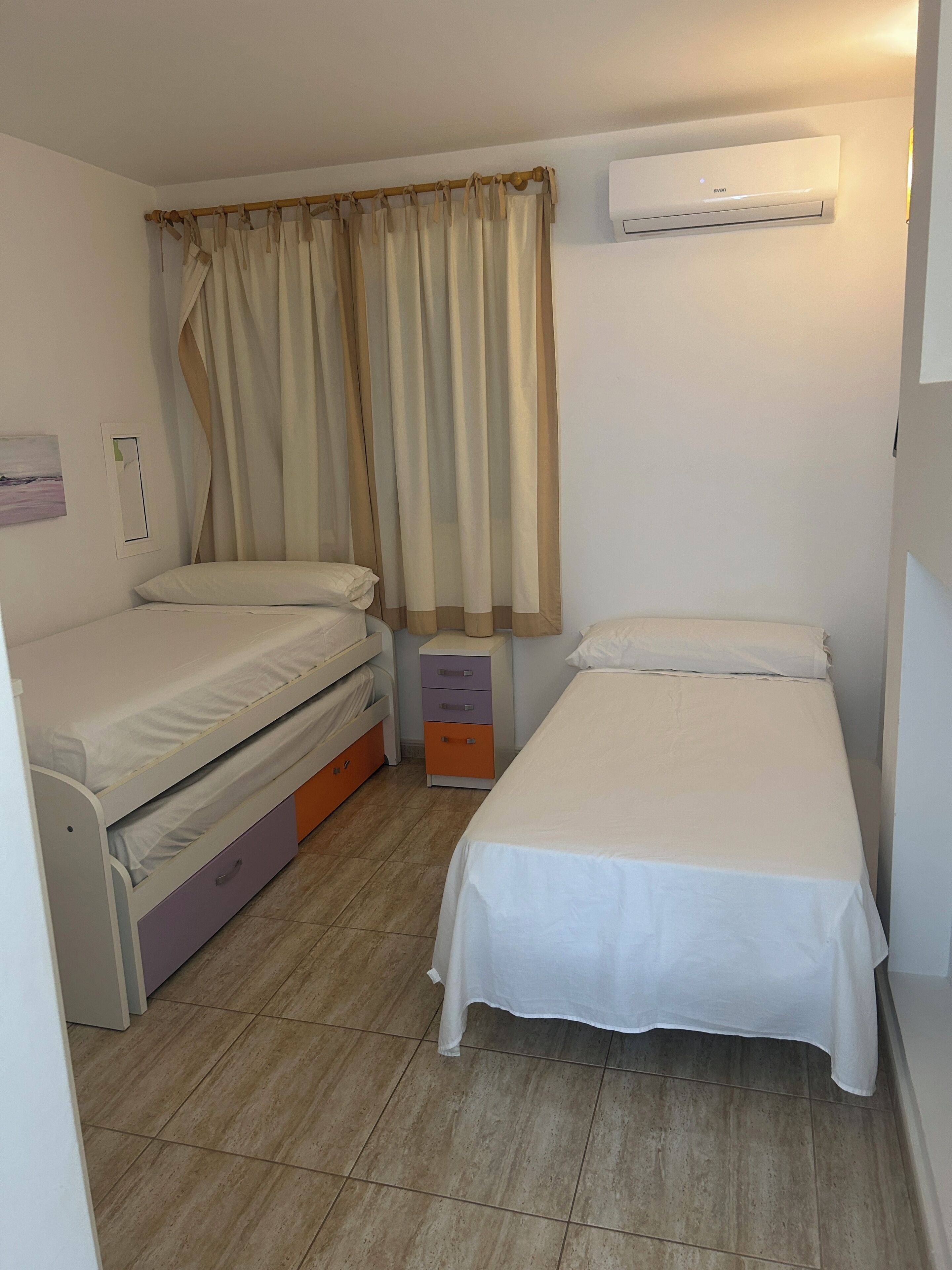 A small, well-lit room with two beds, a wardrobe, a curtain, and an air conditioning unit.
