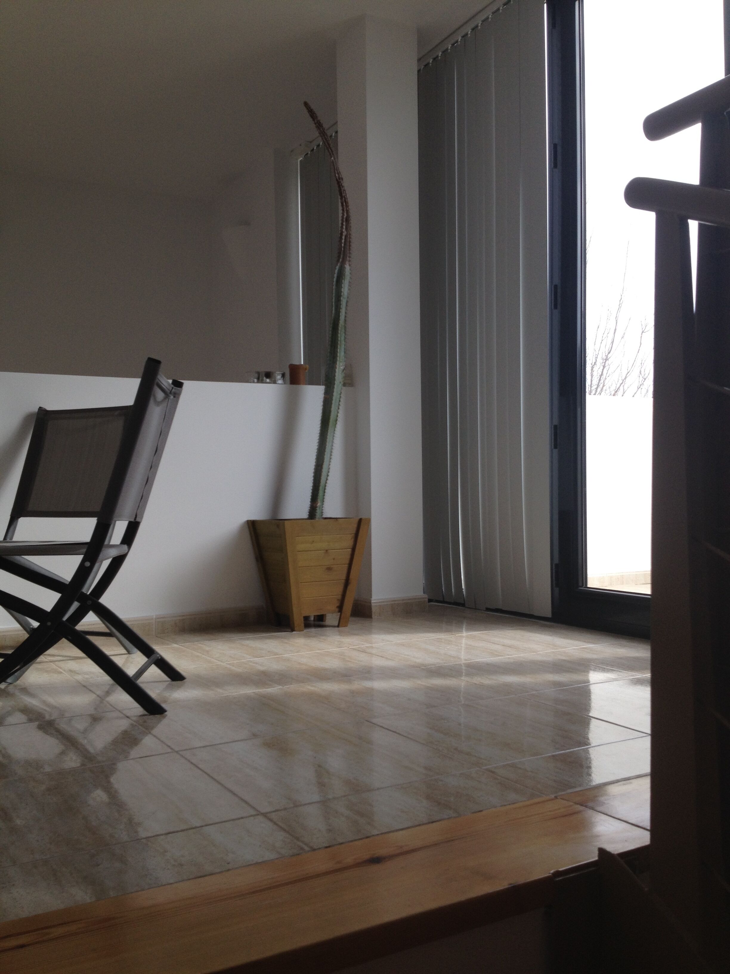Um quarto com uma cadeira de madeira, uma planta alta em vaso de madeira e uma janela grande com cortinas.