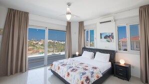 A bedroom with a large bed, a ceiling fan, and a view of the city through large windows.