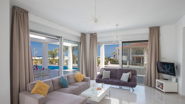 A modern living room with a sofa, coffee table, and TV, large windows with curtains, and a view of the sea.