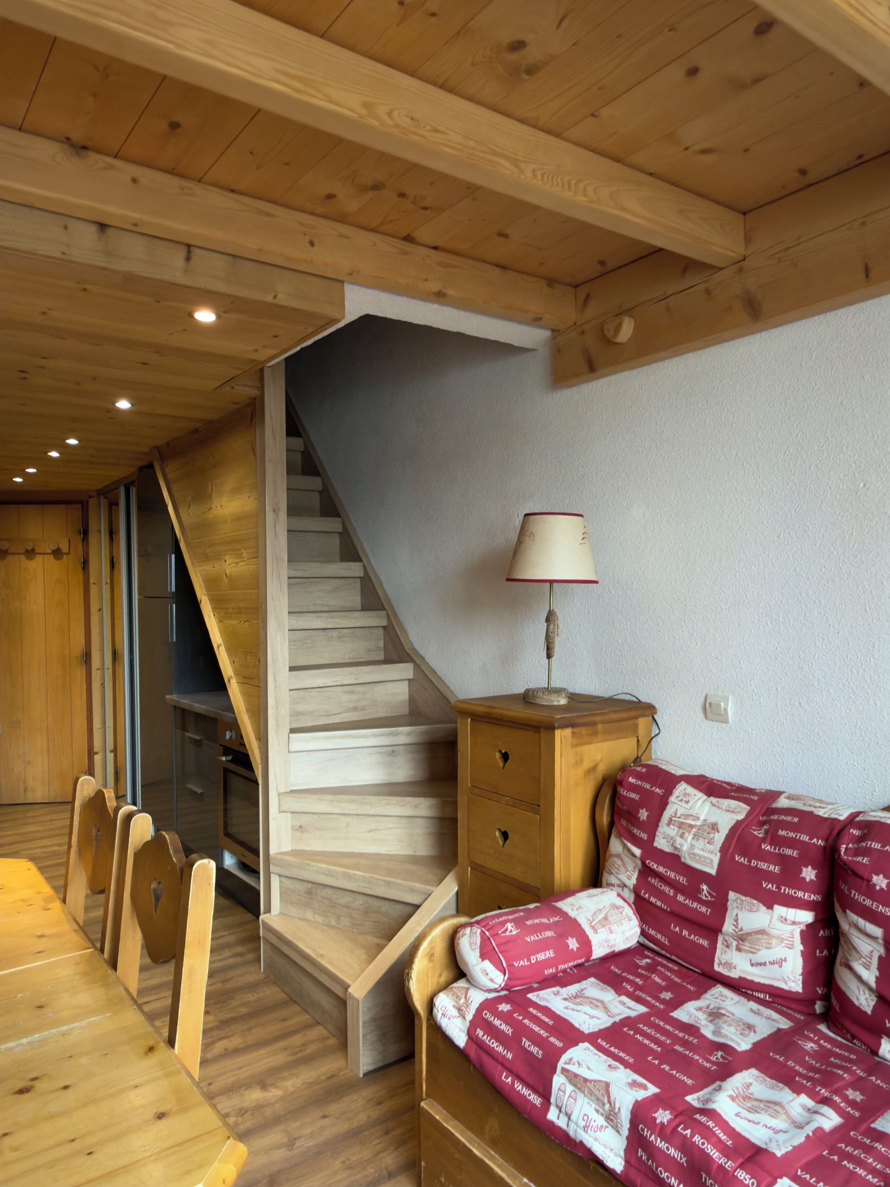 A wooden interior with a staircase, a wooden dining table with chairs, and a sofa with patterned cushions.