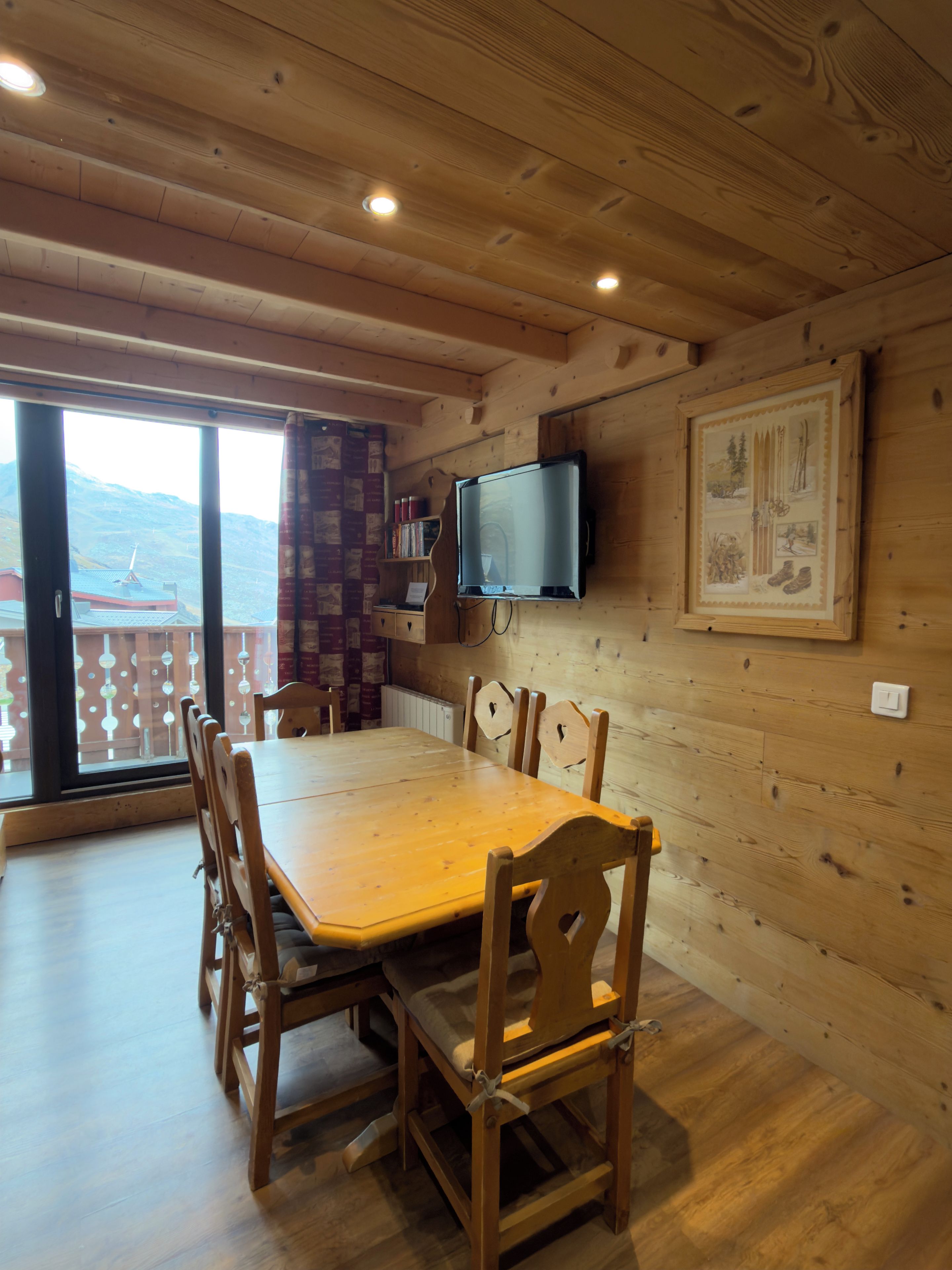 A wooden interior with a dining table, chairs, a mounted television, and a view through a large window.