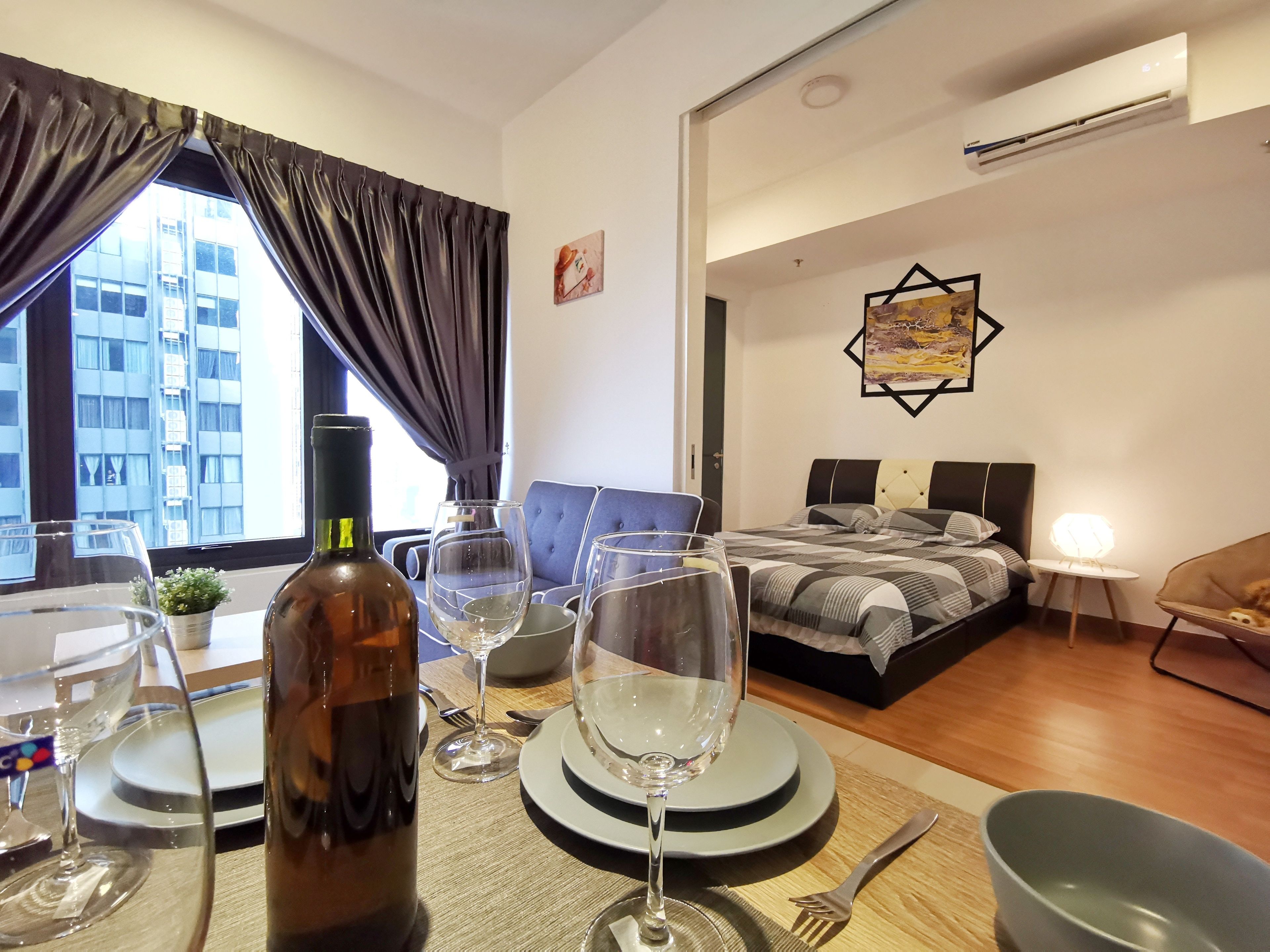 A modern hotel room with a dining area, a bed with a checkered duvet, and a view of a building outside.