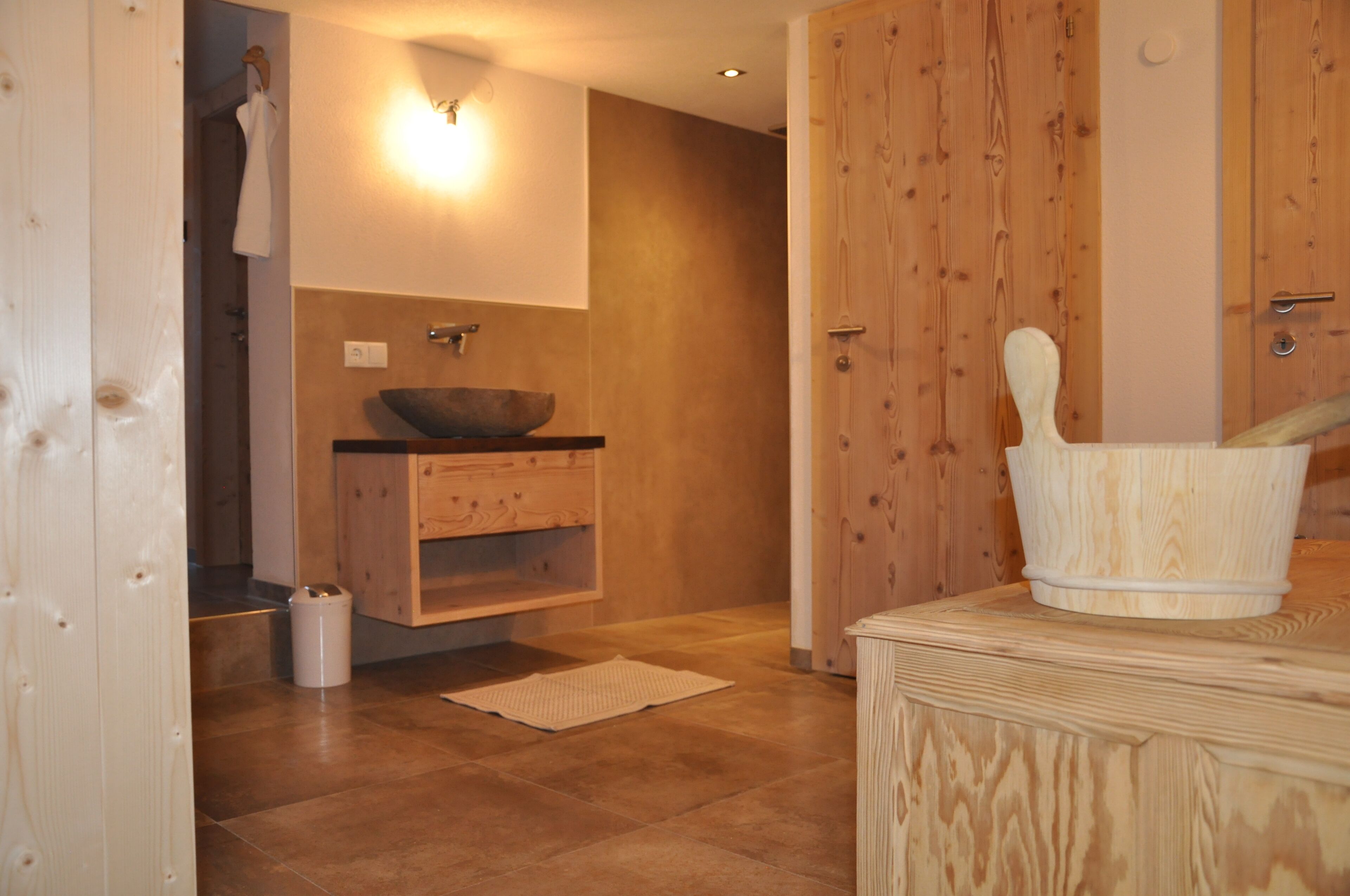 A wooden bathroom with a wooden basin, a wooden cabinet, a white trash bin, and a wooden bucket on a wooden platform.