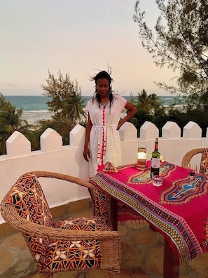 Una persona de pie junto a una mesa con una botella y vasos, con vista a una playa con palmeras y una valla blanca.