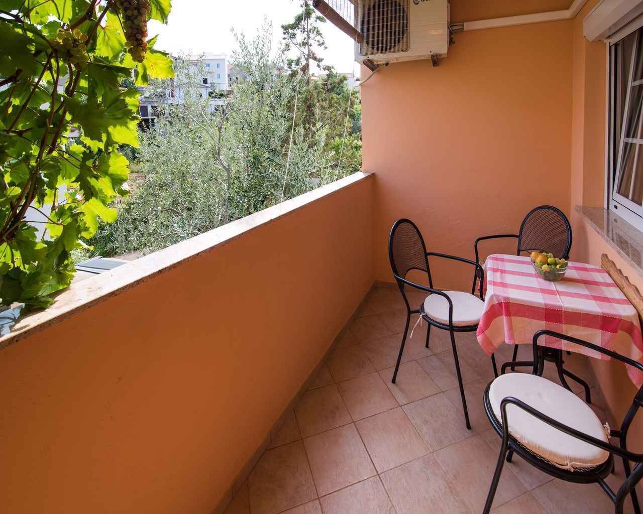 Un balcó amb taula i cadires, una taula petita amb fruita, i vista a arbres i edificis.