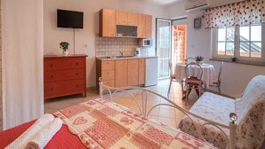 A bedroom with a bed, a bedside table, a red dresser, a small table with chairs, a window with curtains, and a kitchen area with cabinets and a microwave. - Studio with internet access, near the sea (Vrbnik)
