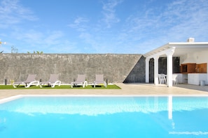 A swimming pool with lounge chairs, a stone wall, and a white building with a kitchen area.