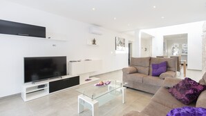 Ett modernt vardagsrum med en platt-TV, ett soffbord i glas, en beige soffa och en matplats i bakgrunden.