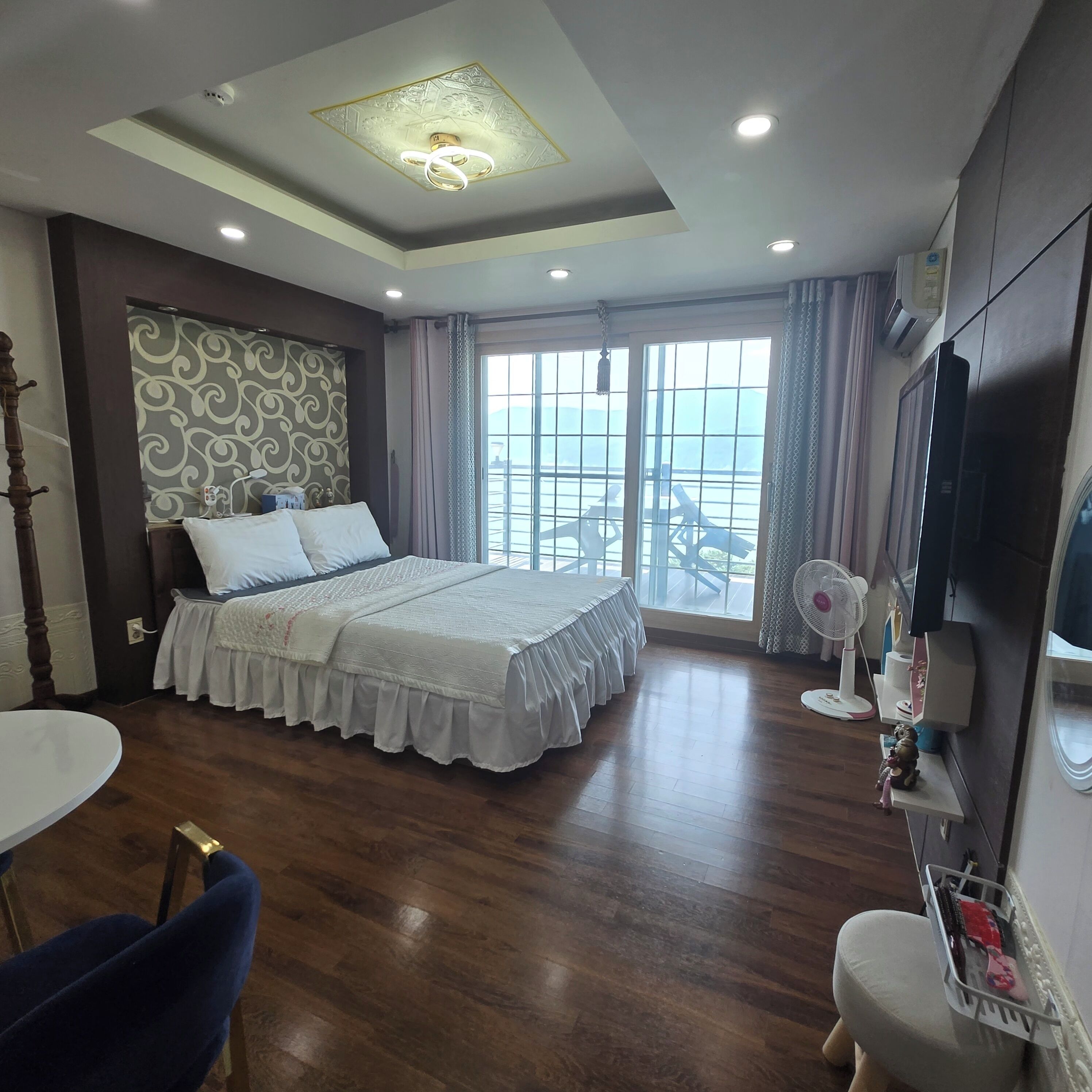 Una habitación de hotel con cama, televisor, ventilador, silla y balcón con vistas.