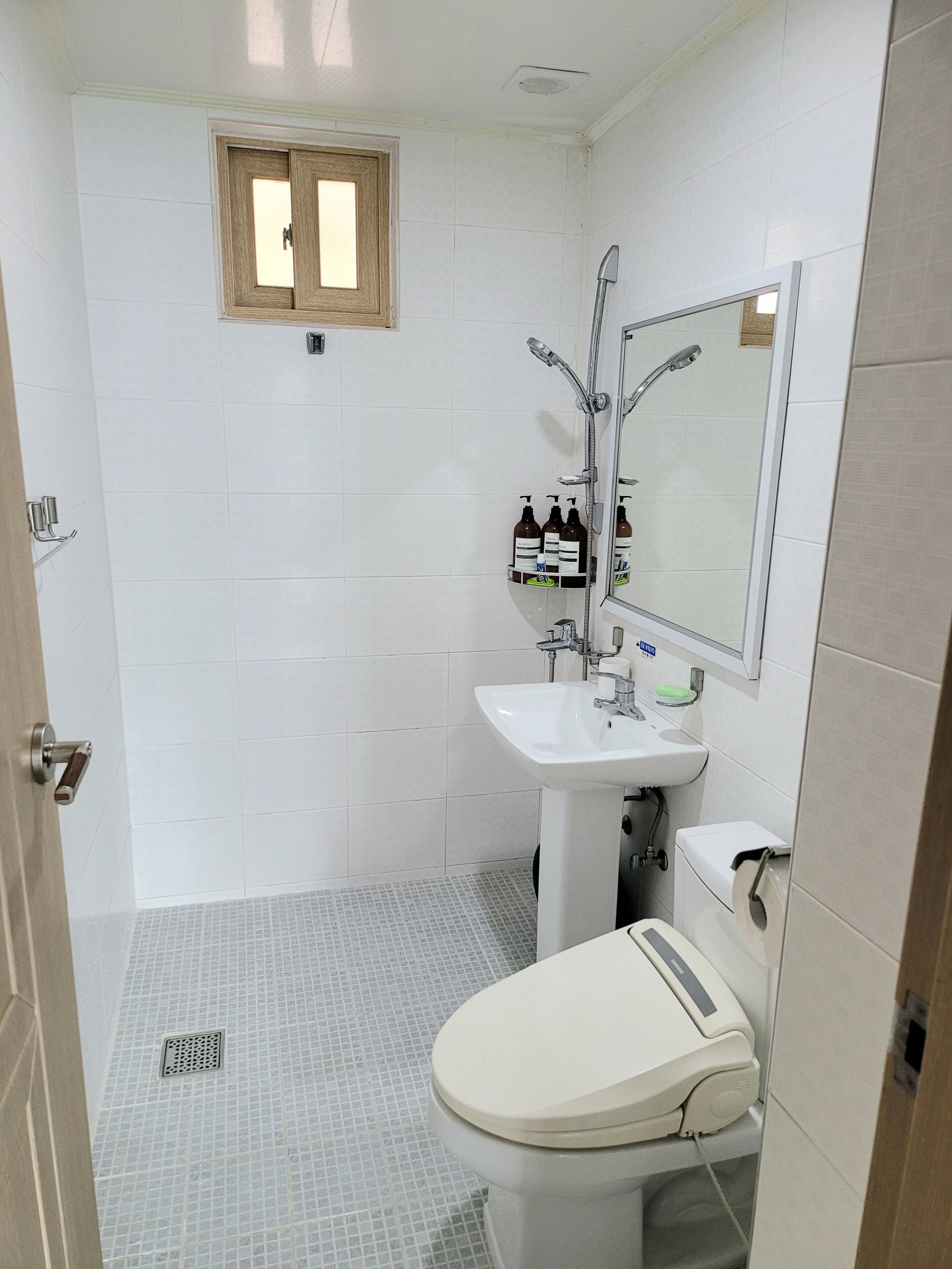 Un baño pequeño y limpio con azulejos blancos, un lavamanos blanco, un inodoro y una ducha. Hay una ventana sobre el lavamanos y un estante con artículos de aseo.
