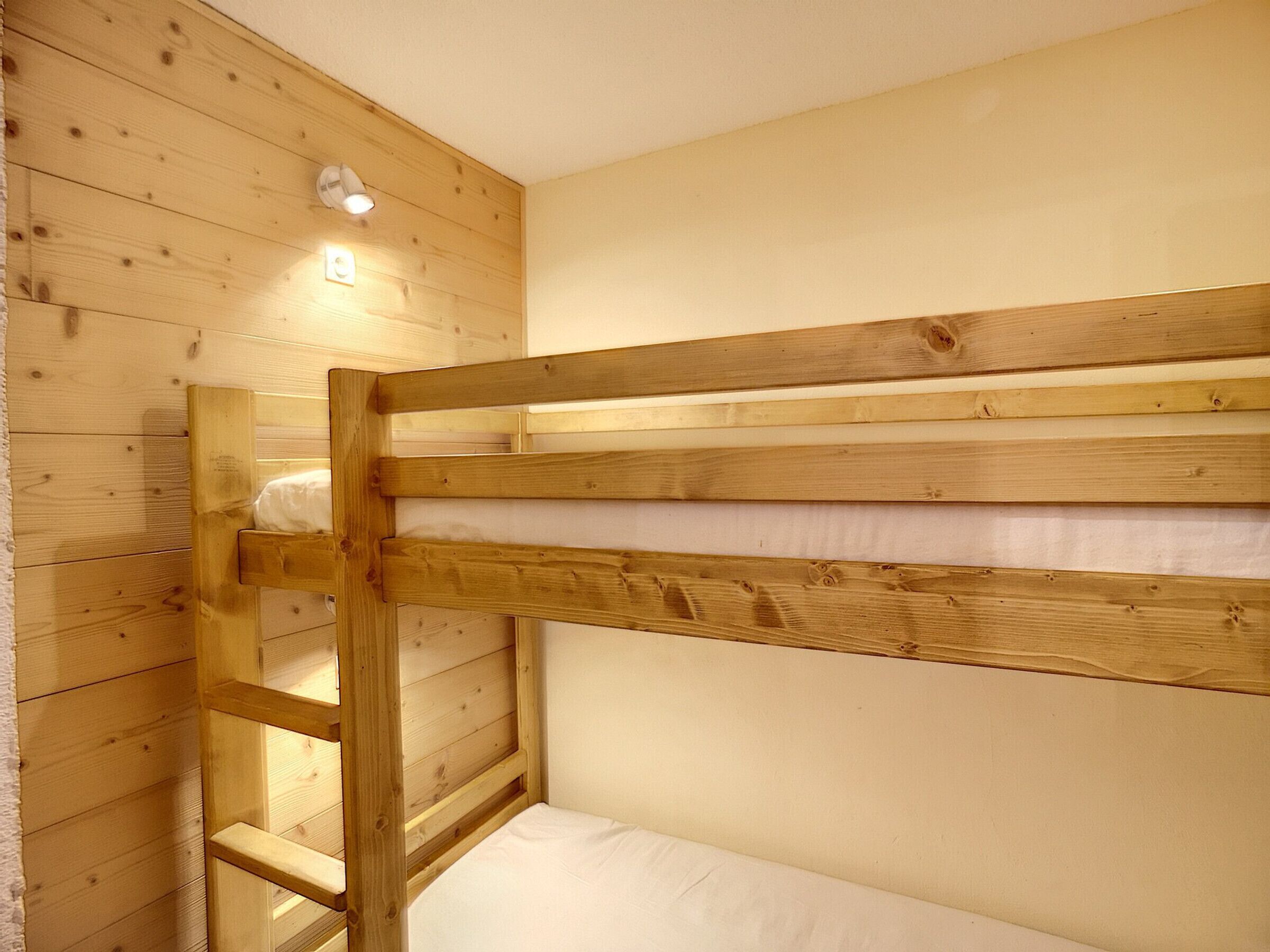 Een stapelbed met houten latten en een witte matras, geplaatst tegen een houten wand met een kleine lamp erboven.