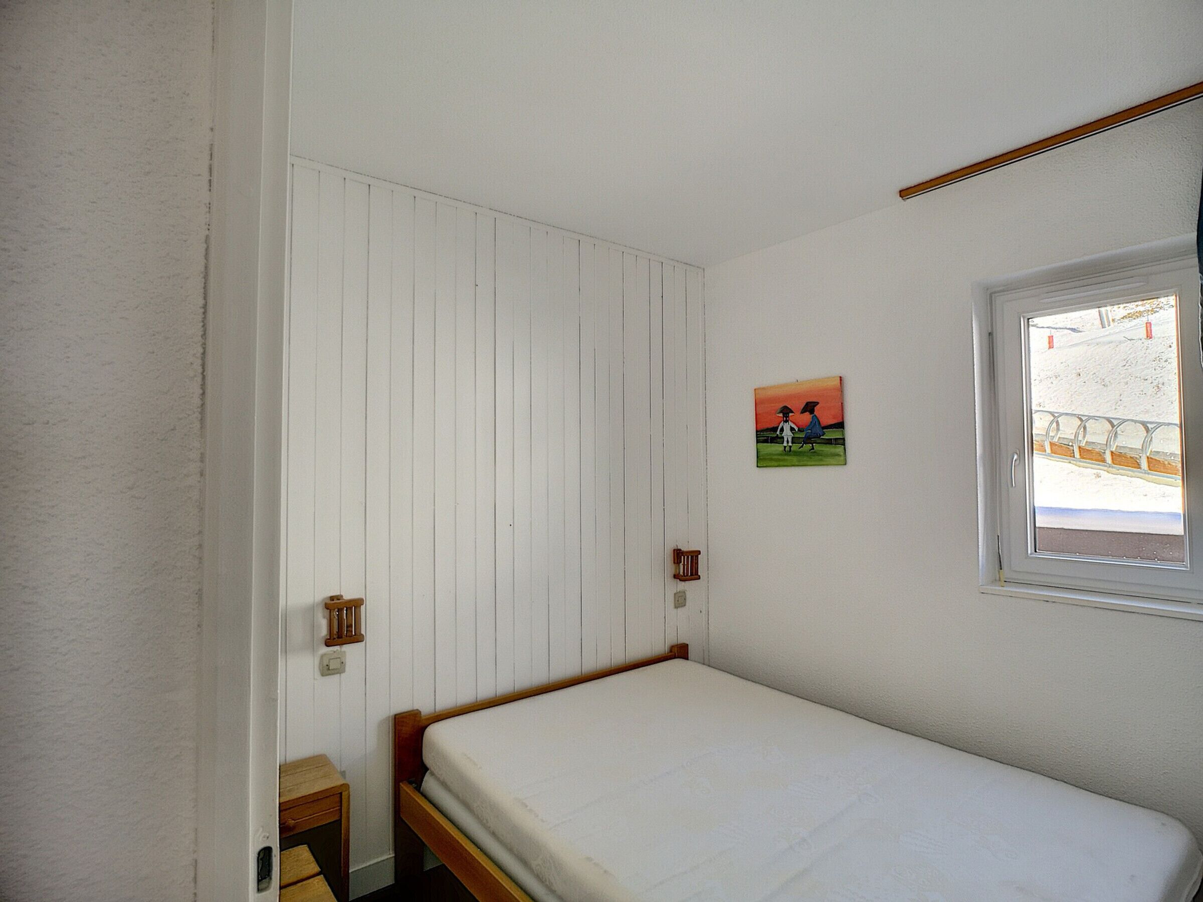 A small, single-bed room with a wooden bed frame, a white wall with vertical paneling, a small bedside table, and a window with a view of a snowy landscape.