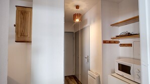 Een smalle gang met houten vloer, witte wanden, een houten kast, een plafondlamp en een magnetron op een witte kast.