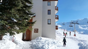 Un immeuble à plusieurs étages, dont le rez-de-chaussée est recouvert de neige, avec un télésiège en arrière-plan et un panneau indiquant un espace public.