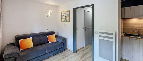 Un appartement moderne avec un canapé gris foncé, un plancher en bois, une cuisine équipée d’un évier et de placards, et une porte donnant sur une autre pièce.
