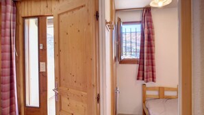 A small, cozy room with a wooden door, a window with a view of a snowy landscape, a bed, and a ceiling light.