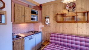 A compact kitchen with wooden cabinets, a built-in microwave, a stove, and a dishwasher. There is a wooden bench with a plaid cushion in the living area.