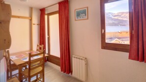 Ein Zimmer mit Holztisch und -stühlen, einem Heizkörper, einer Tür mit roten Vorhängen und einem Fenster mit Landschaftsblick.