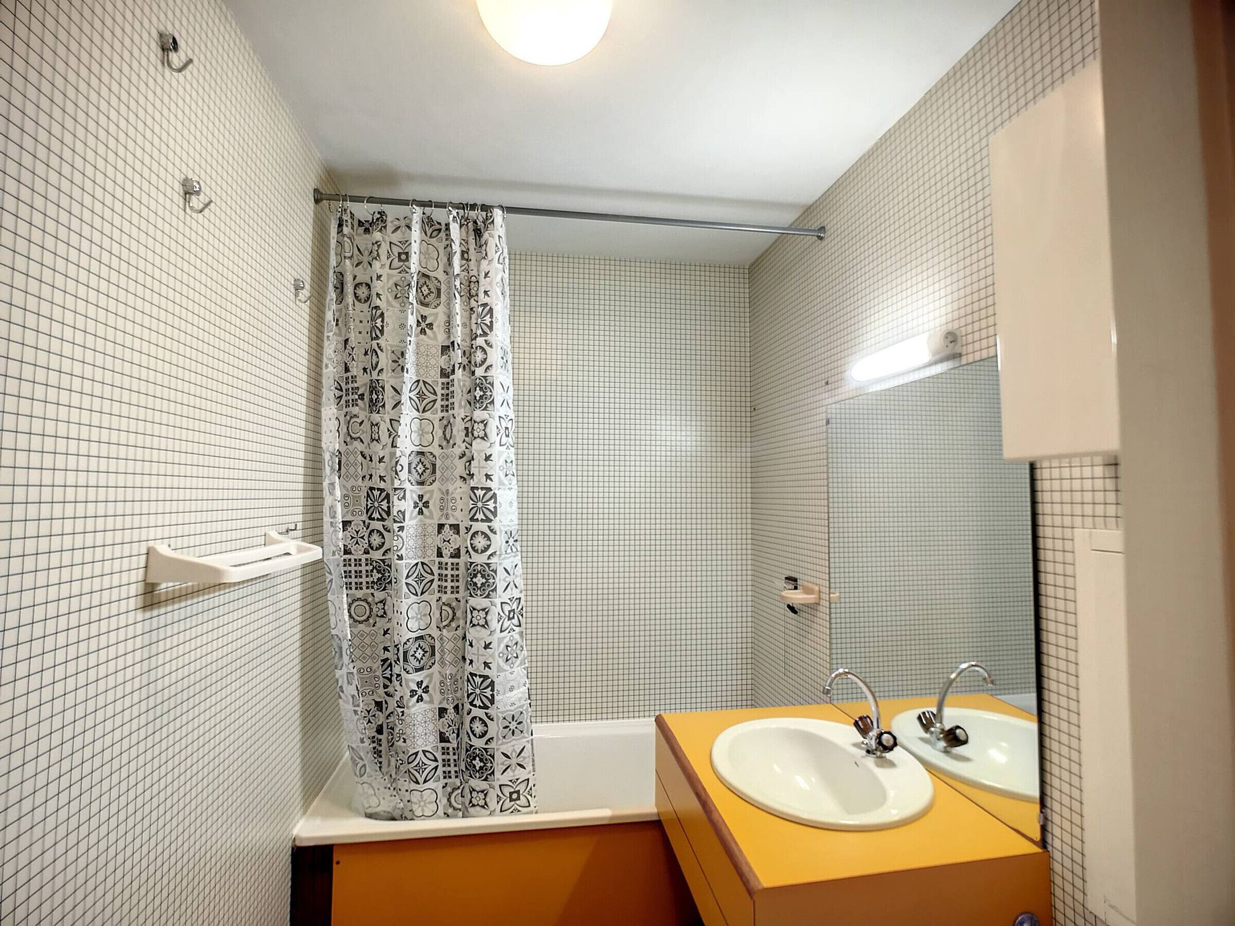 A bathroom with a white tiled wall, a patterned shower curtain, a white sink with a mirror, and a yellow vanity.