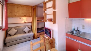 Ein kompakter Wohnraum mit Sofa, Holzregalen, einem kleinen Tisch und einer Küchenzeile mit Spüle und Schränken.