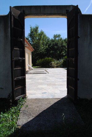 An open gate leading to a courtyard with a tiled floor, surrounded by greenery and a building with a red roof.