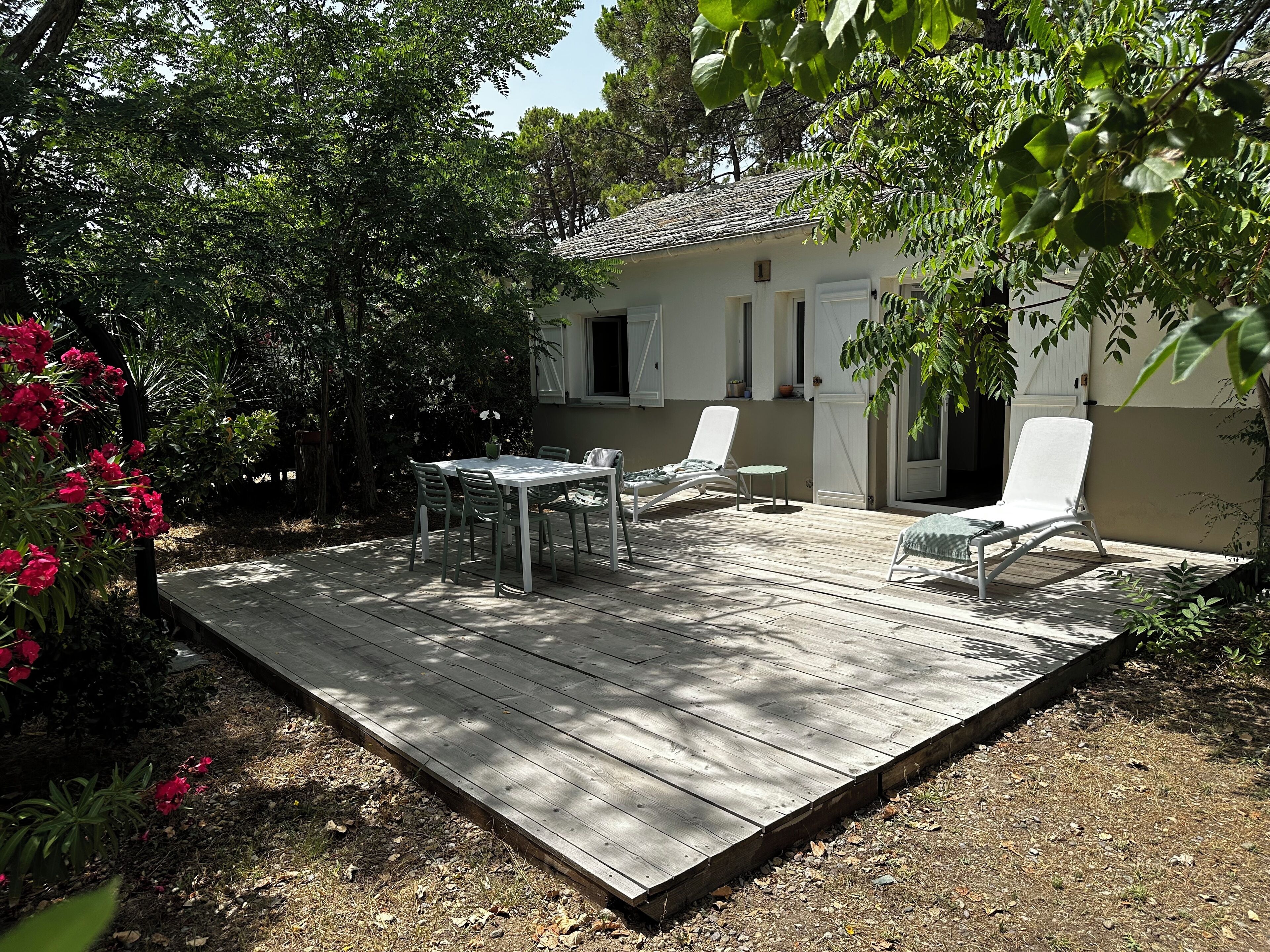 A wooden deck with a table and chairs, surrounded by greenery and a building with white walls and a tiled roof.