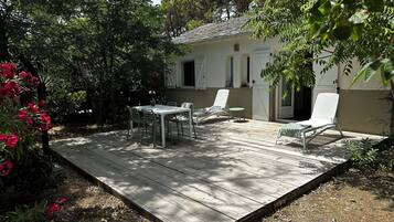 A wooden deck with a table and chairs, surrounded by greenery and a building with white walls and a tiled roof.