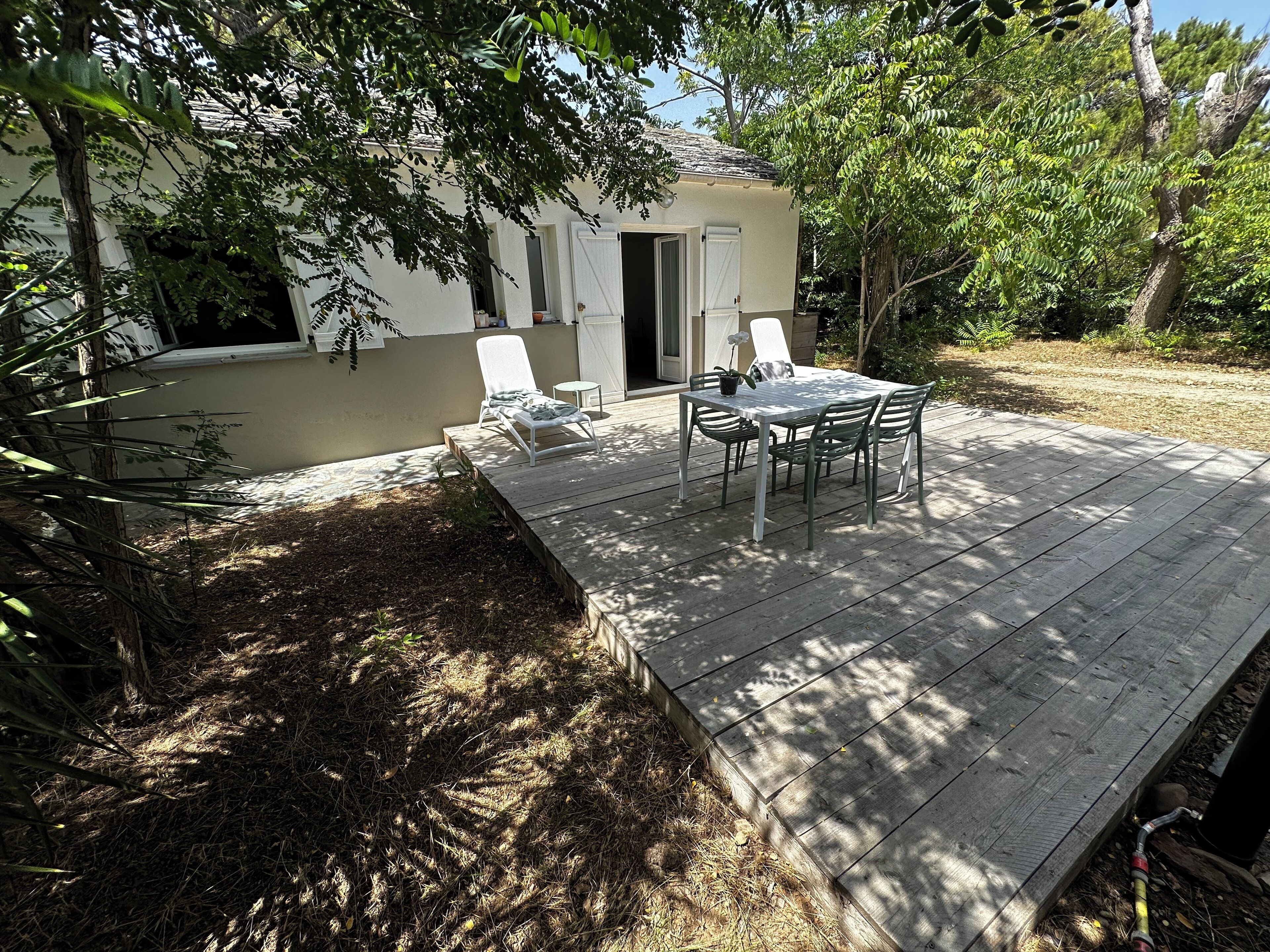 A wooden deck with a table and chairs, surrounded by trees and a building with a door.