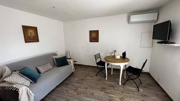 Un salon avec un canapé gris, une table à manger en bois avec des chaises, un téléviseur fixé au mur et deux tableaux encadrés accrochés au mur.