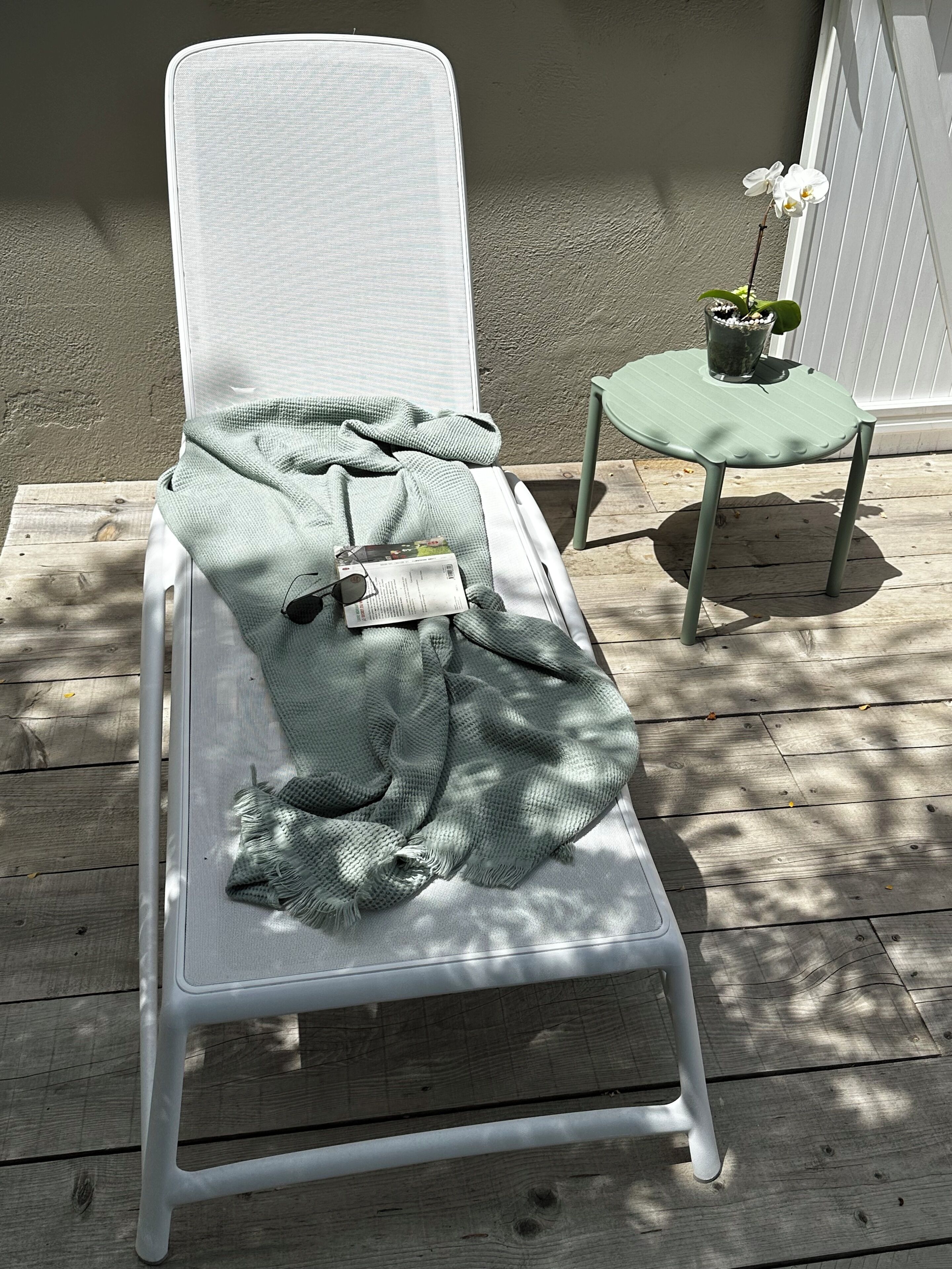 Una silla lounge blanca con una manta verde y gafas de sol, sobre una terraza de madera.