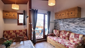 A cozy room with a wooden ceiling, a sofa with a quilted cover, a dining table with chairs, and a window with curtains.