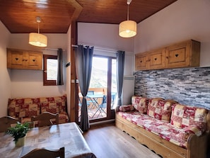 Um quarto acolhedor com teto de madeira, um sofá com manta acolchoada, uma mesa de jantar com cadeiras e uma janela com cortinas.