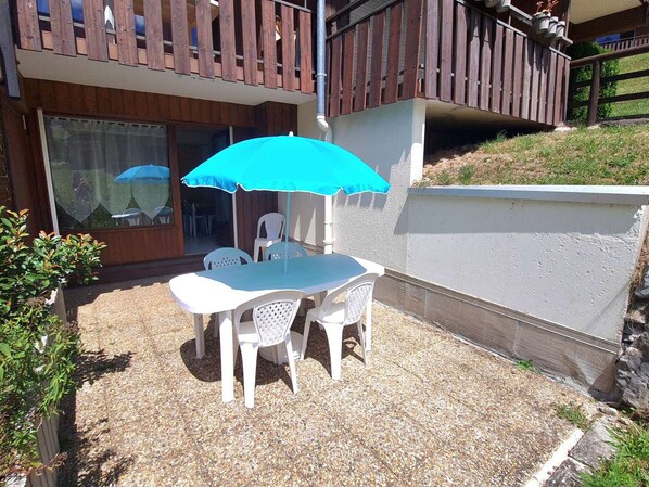 A patio with a table and chairs under a blue umbrella, a wooden deck, and a white wall.