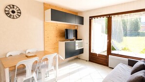 A compact living space with a kitchenette, a dining area with a wooden table and white chairs, a sofa, and a large window with a view of greenery.