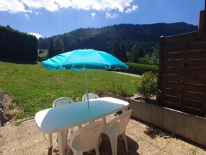 An outdoor patio with a table and chairs, a blue umbrella, and a view of a forested hill.