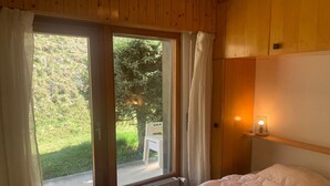 A bedroom with a bed, a window with curtains, a small table, and a nightstand with a lamp.