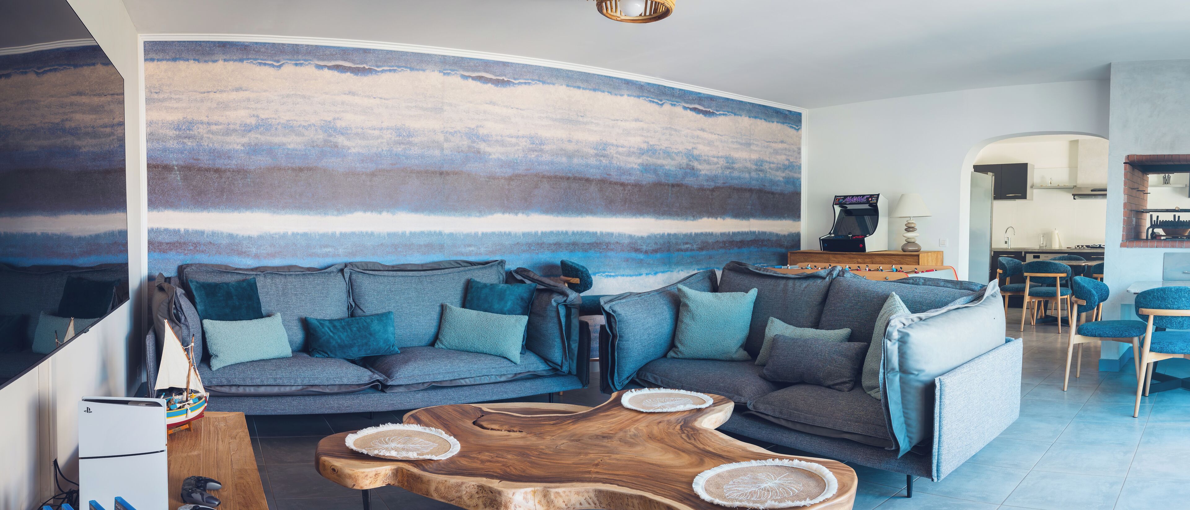 A living room with a curved wall mural, a gray sofa with teal cushions, a wooden coffee table, and a kitchen area in the background.