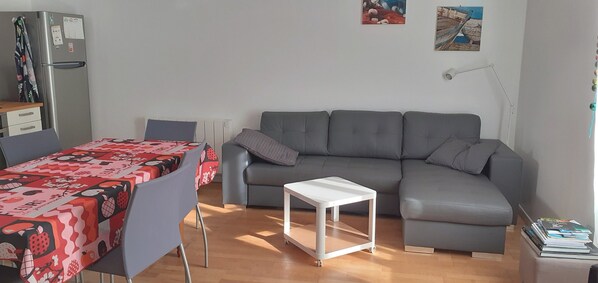 A modern living room with a grey sofa, a small white table, a dining table with a red and black patterned tablecloth, a refrigerator, and a lamp. - Holiday house 6 pers. in Etel, beach at 100 m (Étel)