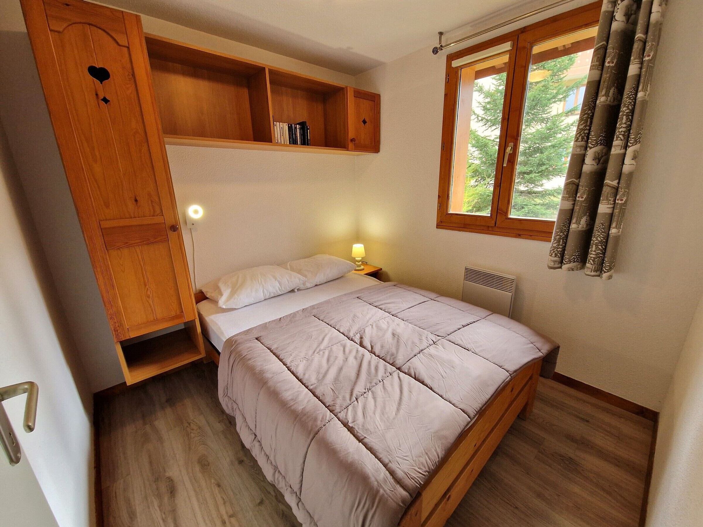 A bedroom with a bed, wooden shelves, a nightstand, a window with curtains, and a radiator.