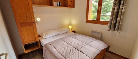 A bedroom with a bed, wooden shelves, a nightstand, a window with curtains, and a radiator.