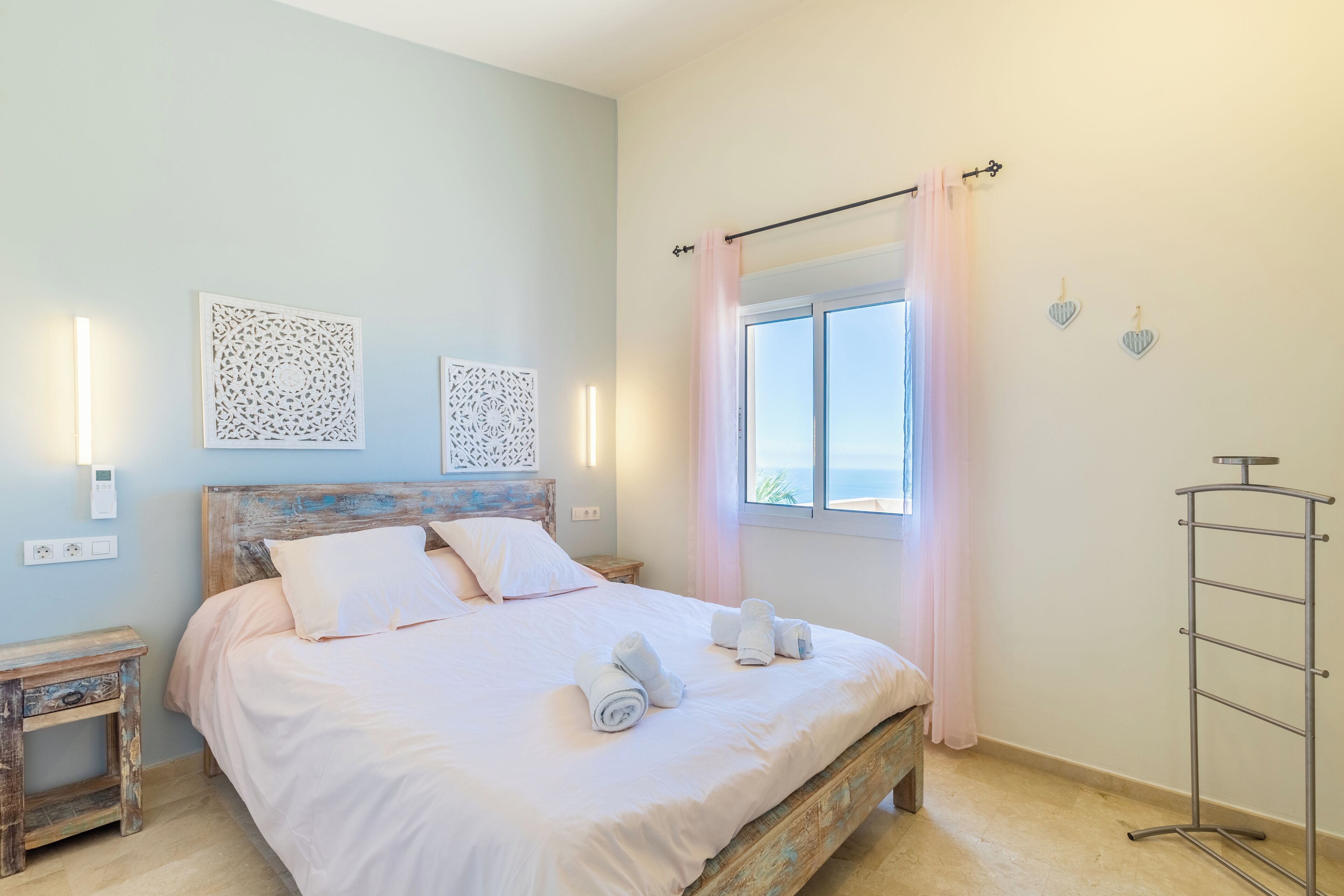 Una cama bien hecha con almohadas y toallas enrolladas, una mesita de noche, un aplique de pared y una ventana con cortinas.
